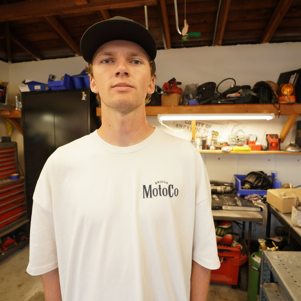 Dayton wearing a cream colored motocross tshirt with a chest print of Briggs MotoCo in navy and grey in a workshop
