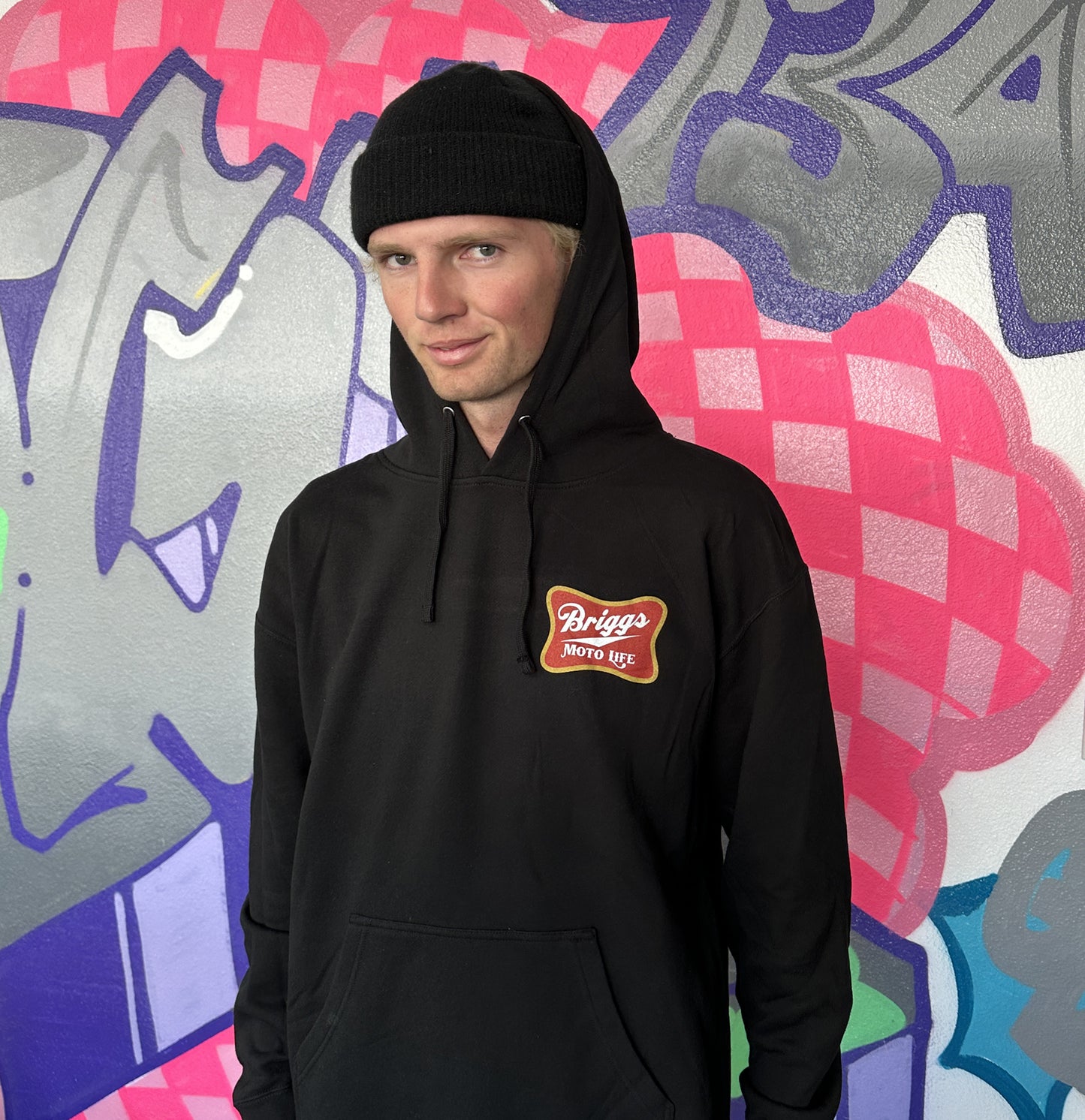 Dayton wearing a black hoodie with 'Briggs Moto Life' logo, spoofing the old school 'Miller High life' badge logo on the front pocket area, standing in front of a colorful graffiti wall.