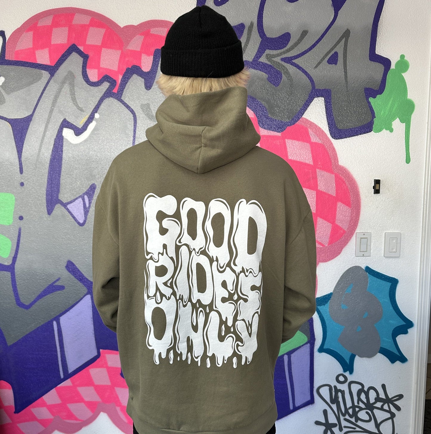 Dayton is wearing an olive green hoodie with 'GOOD RIDES ONLY' printed in white on the back, standing in front of a graffiti wall.