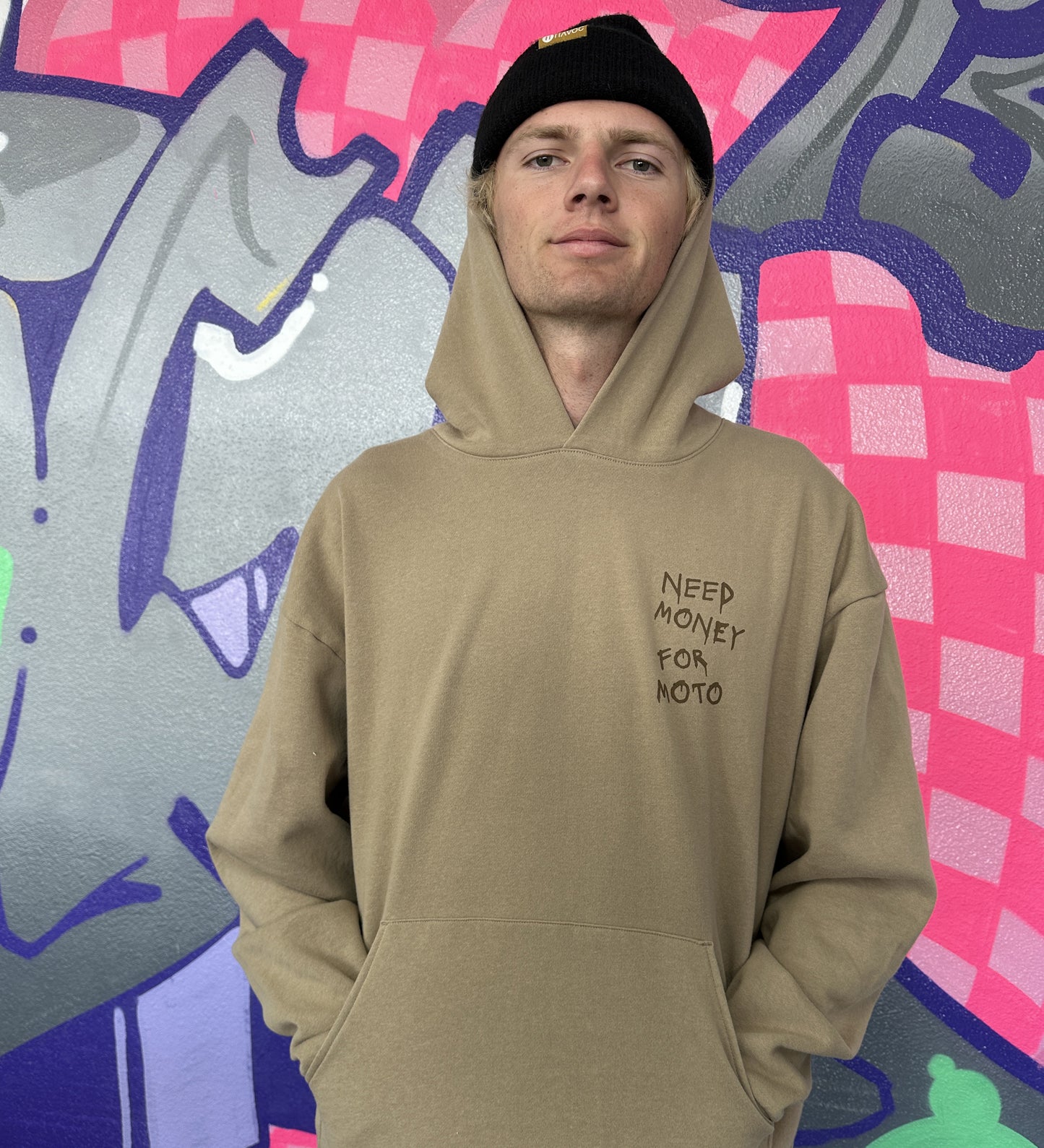 Dayton is wearing a tan hoodie with brown text 'NEED MONEY FOR MOTO' printed in a spray-paint like font on the left chest area, standing in front of a colorful graffiti wall.