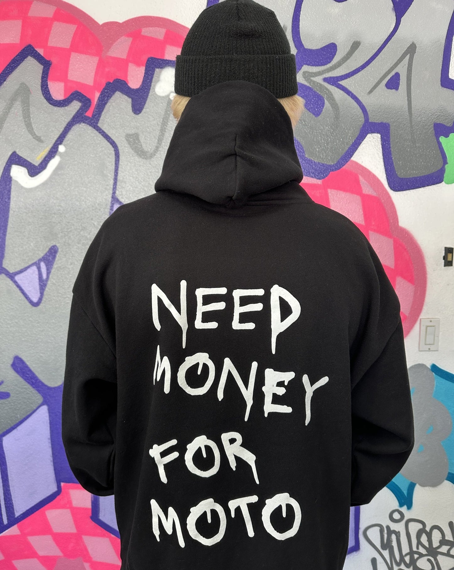 Dayton wearing a black hoodie with the text 'NEED MONEY FOR MOTO' printed in white on the back, standing in front of a graffiti wall.