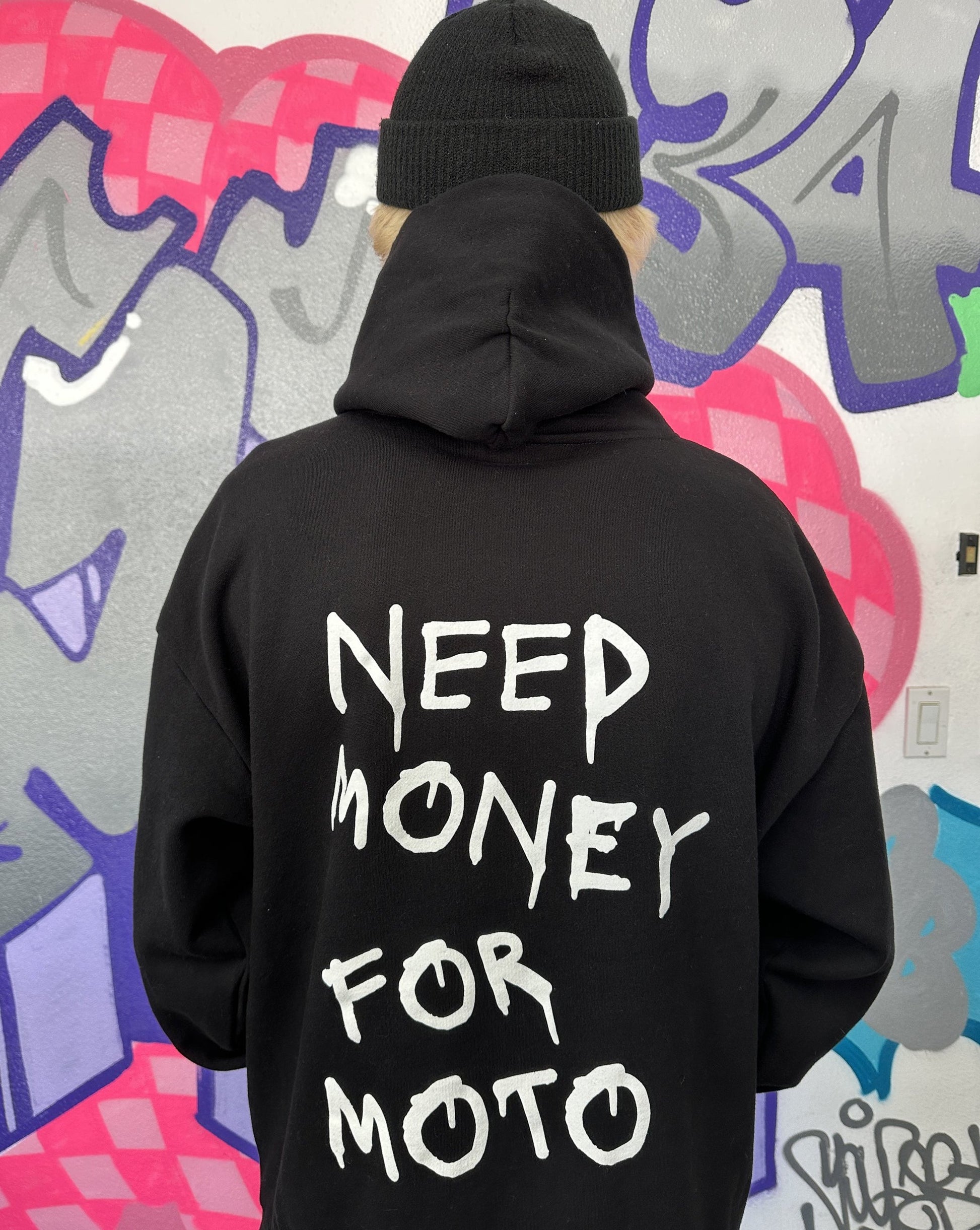 Dayton wearing a black hoodie with the text 'NEED MONEY FOR MOTO' printed in white on the back, standing in front of a graffiti wall.