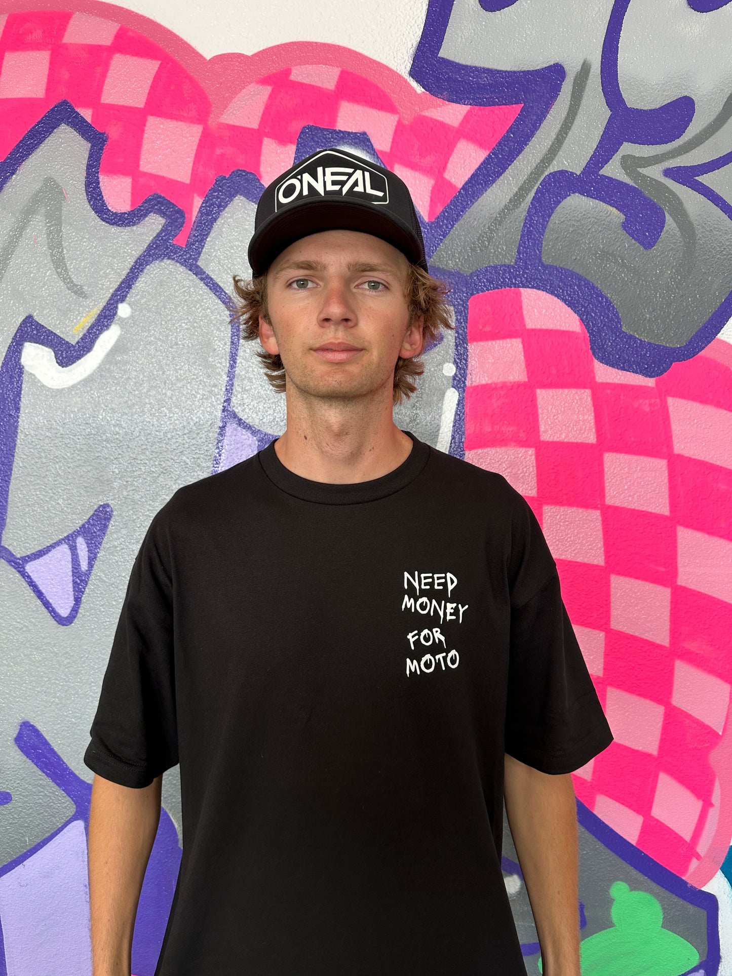 Dayton wearing a black short sleeve T-shirt with white text 'Need Money For Moto' printed on the front and back, standing in front of a wall with colorful graffiti.