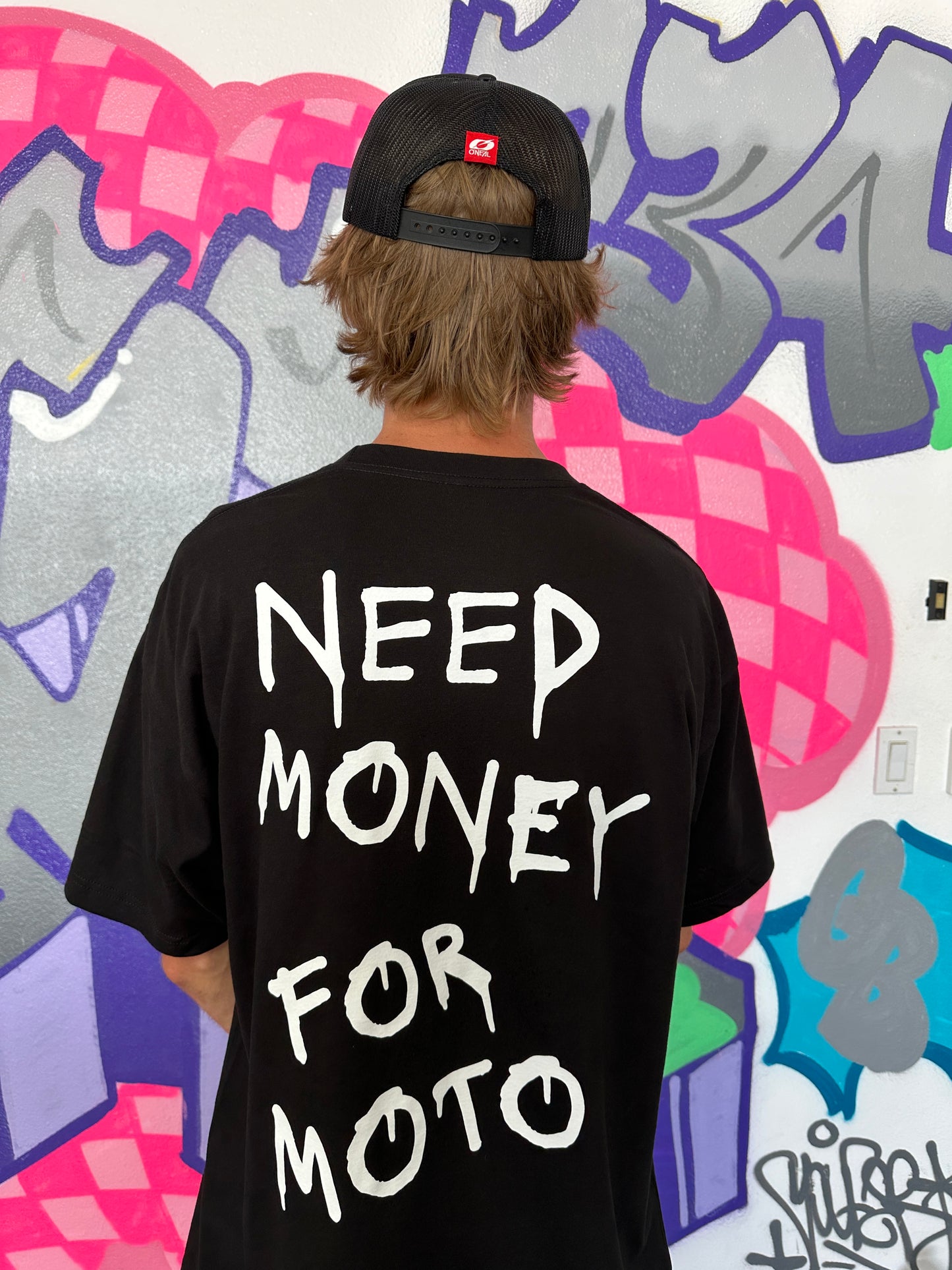 Dayton wearing the black tshirt that says 'Need Money For Moto' shirt with white printing in front of a graffiti wall.