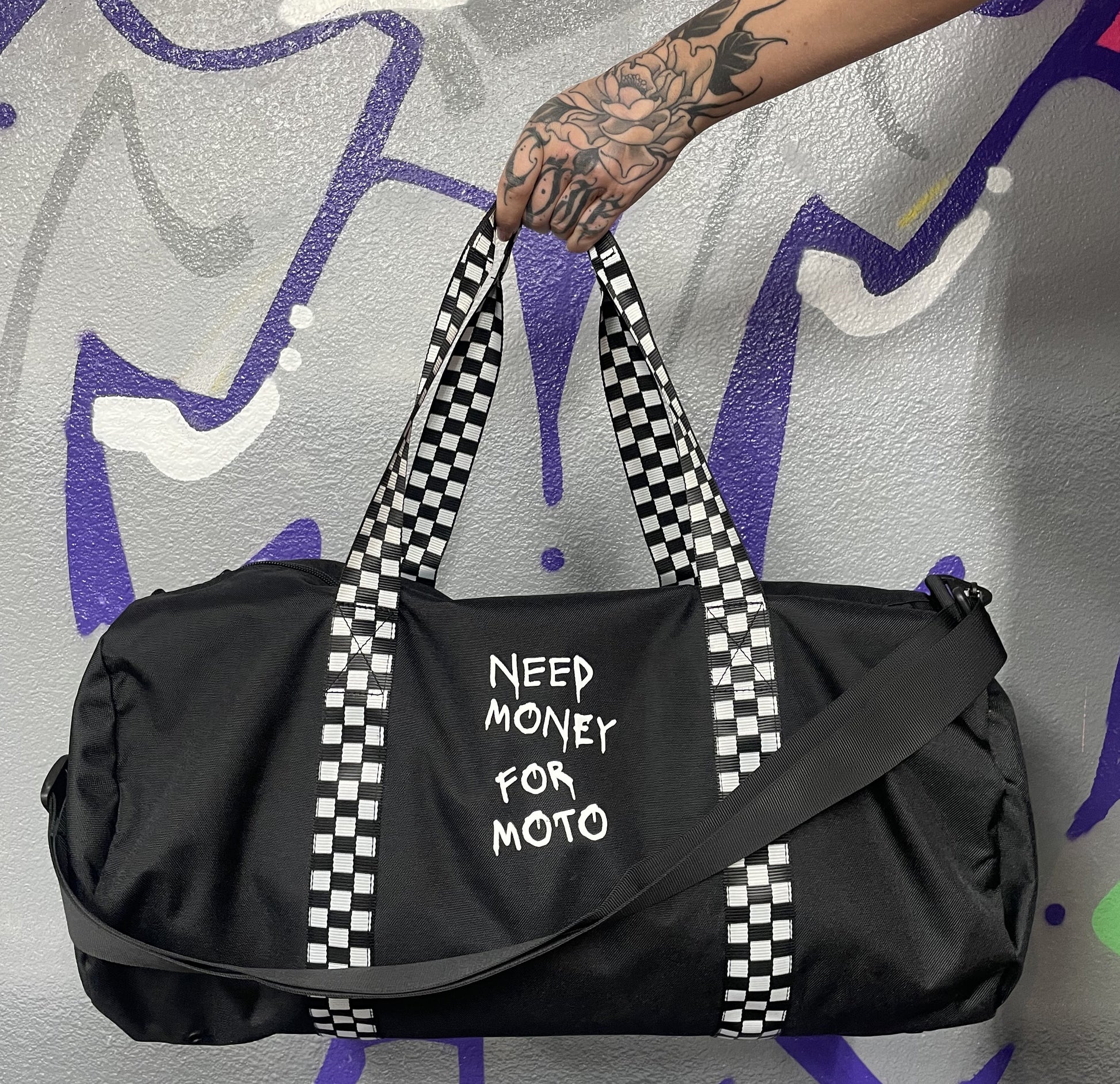 A black duffel bag with checkered handles and the text 'NEED MONEY FOR Moto' printed on the side. The bag is displayed against a graffiti background.
