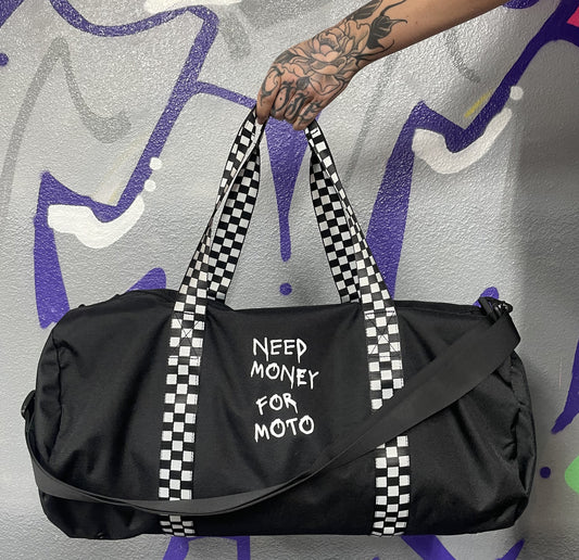 A black duffel bag with checkered handles and the text 'NEED MONEY FOR Moto' printed on the side. The bag is displayed against a graffiti background.